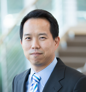 A person wearing a dark suit and blue striped tie is smiling slightly. The background is softly blurred, showing hints of a modern interior with glass and stairs.
