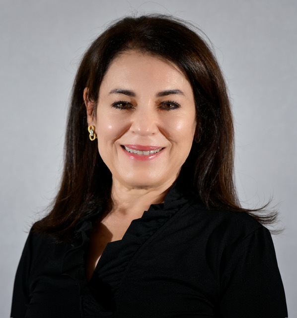 Woman with shoulder-length dark brown hair, wearing a black top, smiles at the camera against a plain, light-colored background.