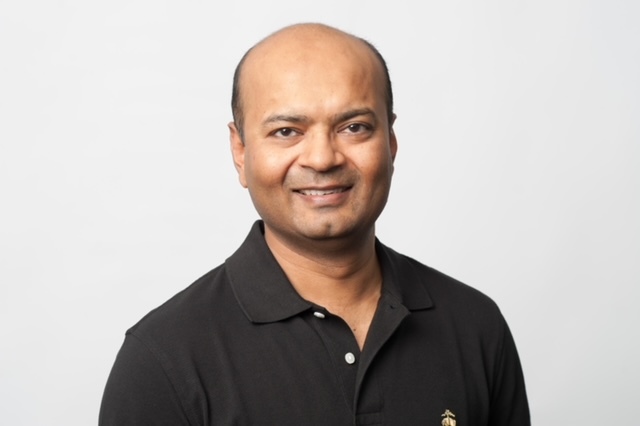 A person with a shaved head and a slight smile is wearing a black collared shirt against a plain white background.
