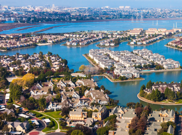 Aerial view of a suburban neighborhood with houses, tree-lined streets, and waterways, set against a backdrop of a large city and a body of water in the distance.
